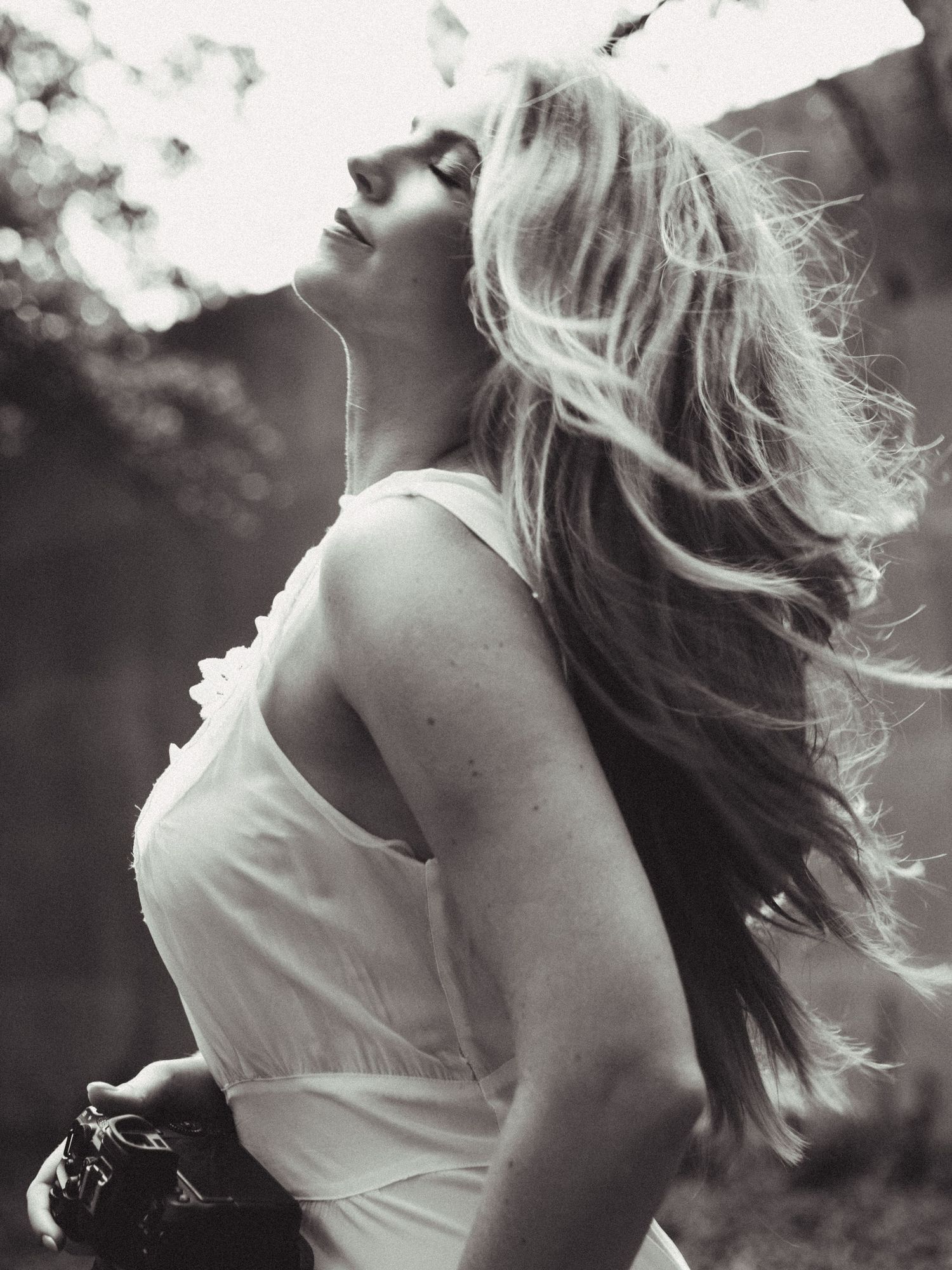 Black and white photo of a blonde woman in a white dress with flowing hair, gazing upward outdoors.