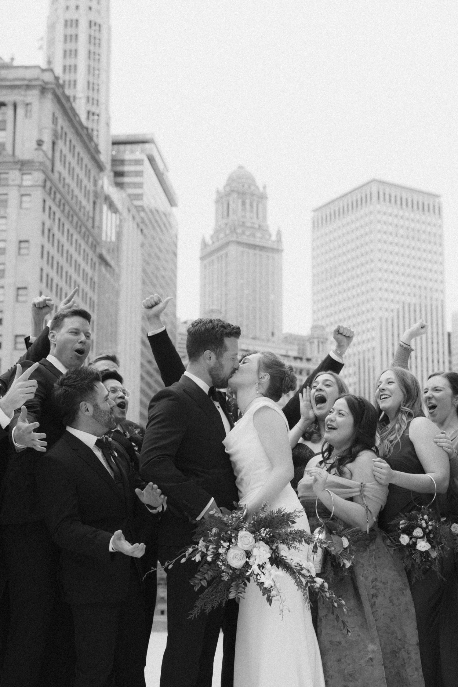 Wedding party celebrates joyfully against urban backdrop of tall buildings in black and white photo series.