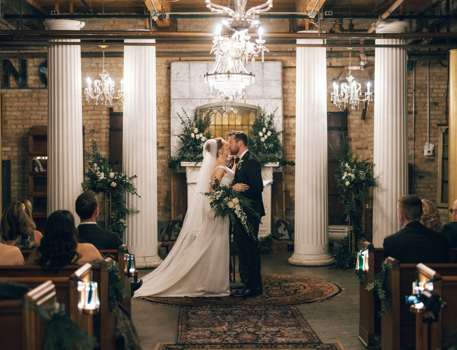 A wedding ceremony takes place in an elegant venue with white columns, chandeliers, and stone walls creating a romantic atmosphere.