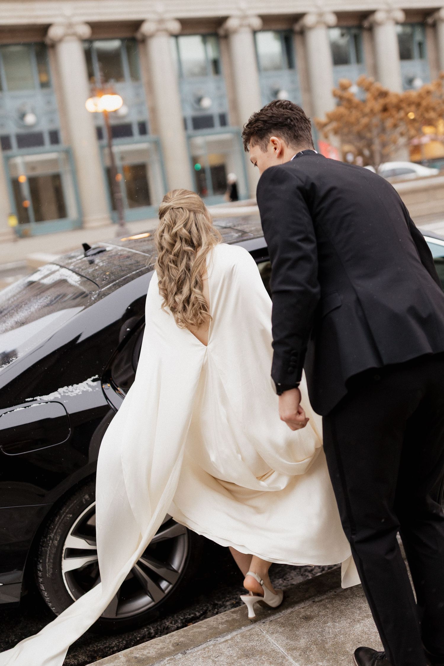 A couple in wedding attire enters a black car outside a classical building.