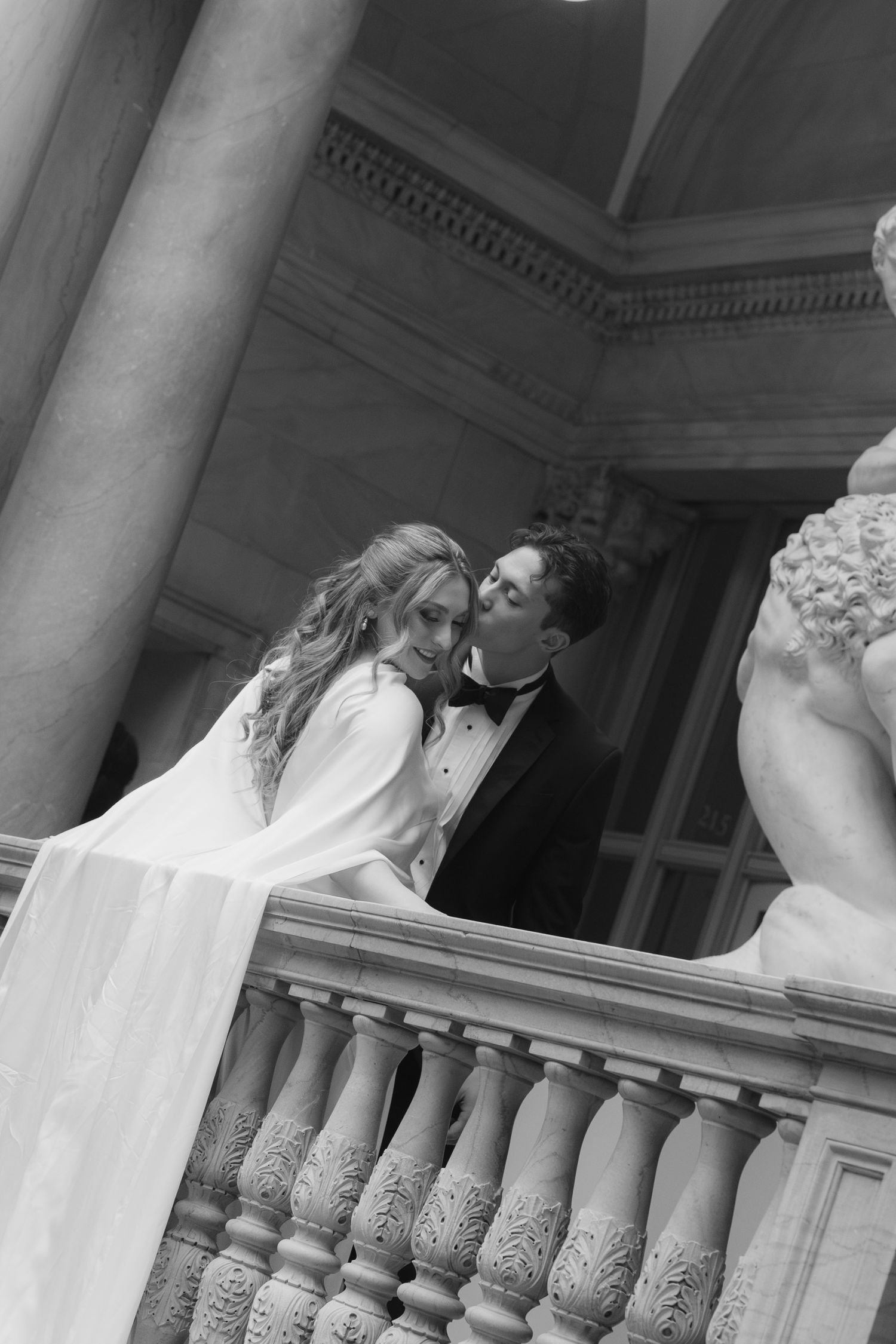 A romantic wedding couple shares an intimate moment on an elegant stone balcony with ornate balusters.
