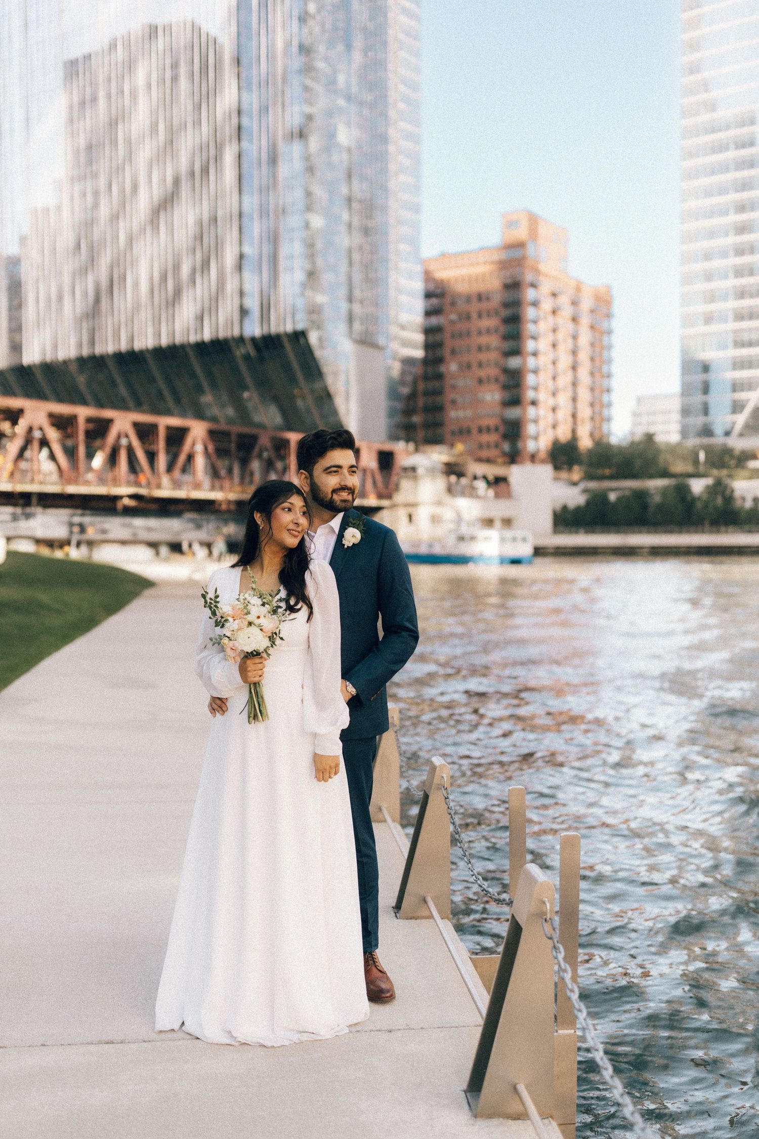 A couple in wedding attire embraces by the waterfront with city buildings in the background.