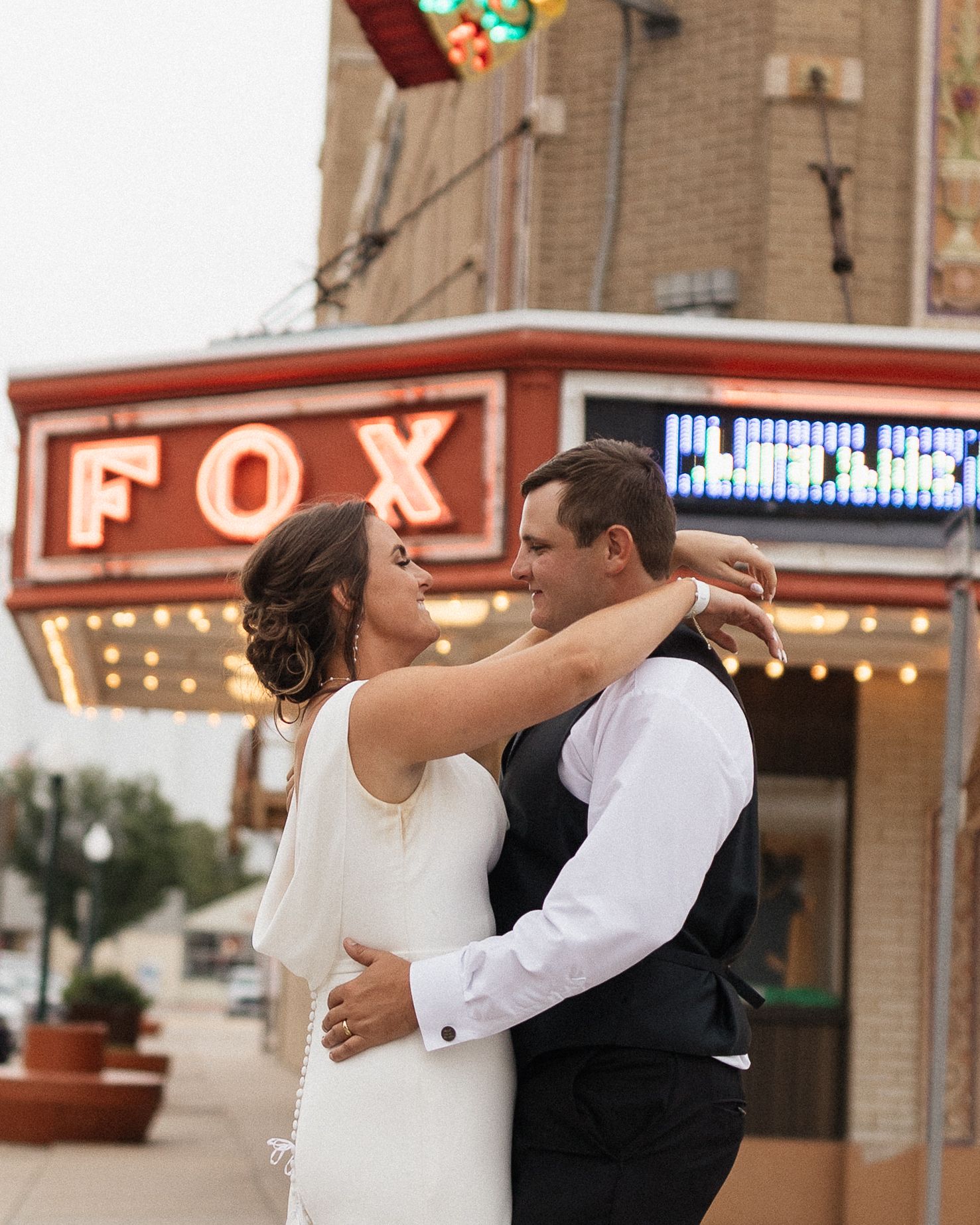 A couple in wedding attire dances and embraces outside a historic brick building with theater marquee lighting.