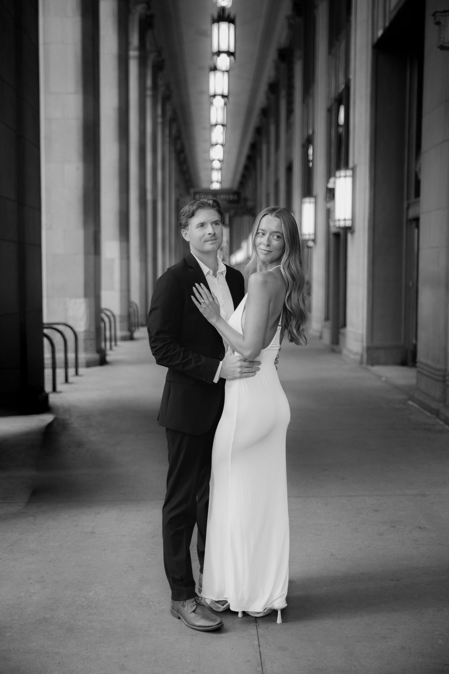 Black and white photo of a couple embracing in an elegant columned hallway, woman in white gown, man in dark suit.