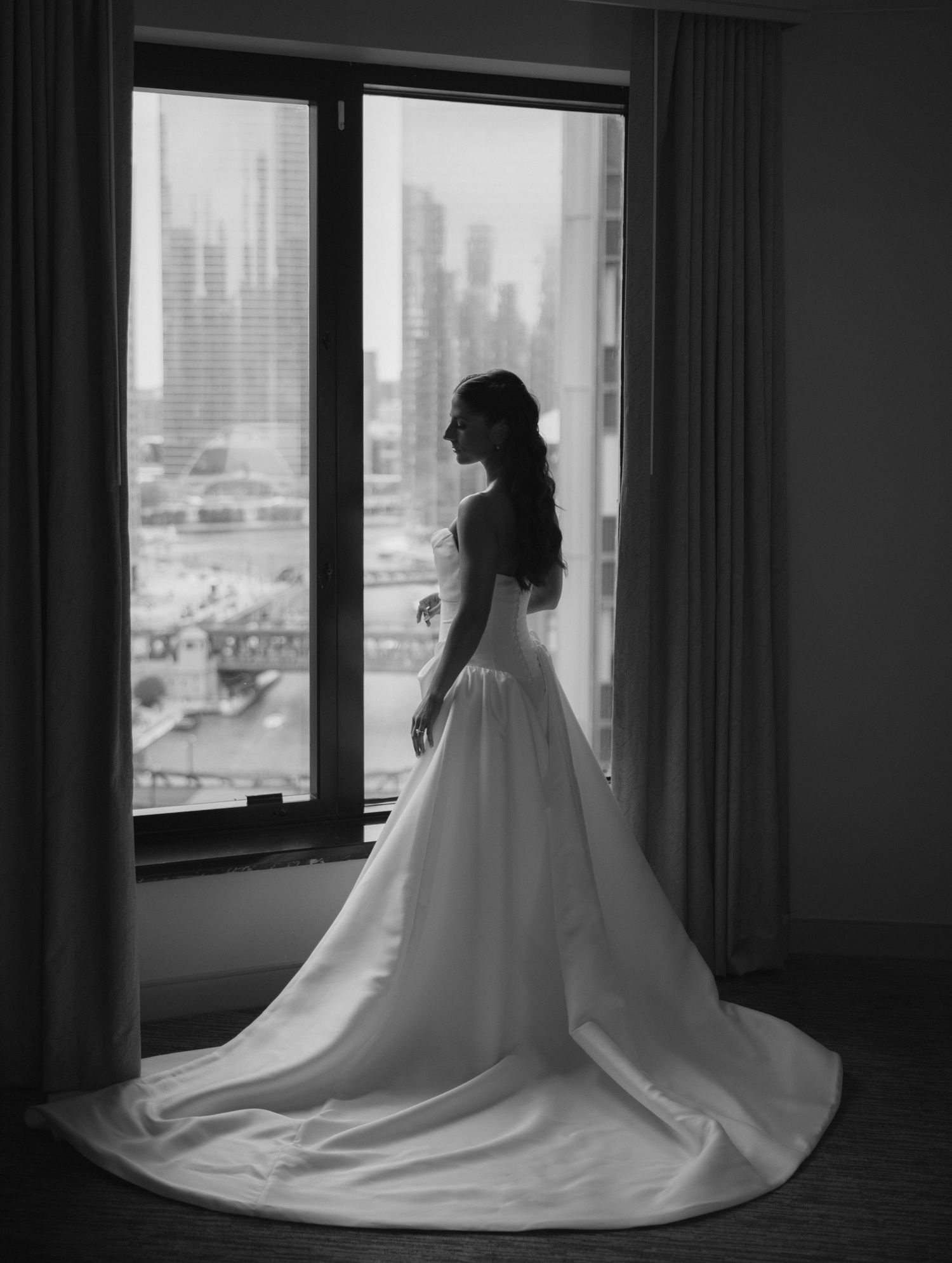 Bride in elegant white ball gown with long train stands by large window overlooking city skyline in black and white photo.