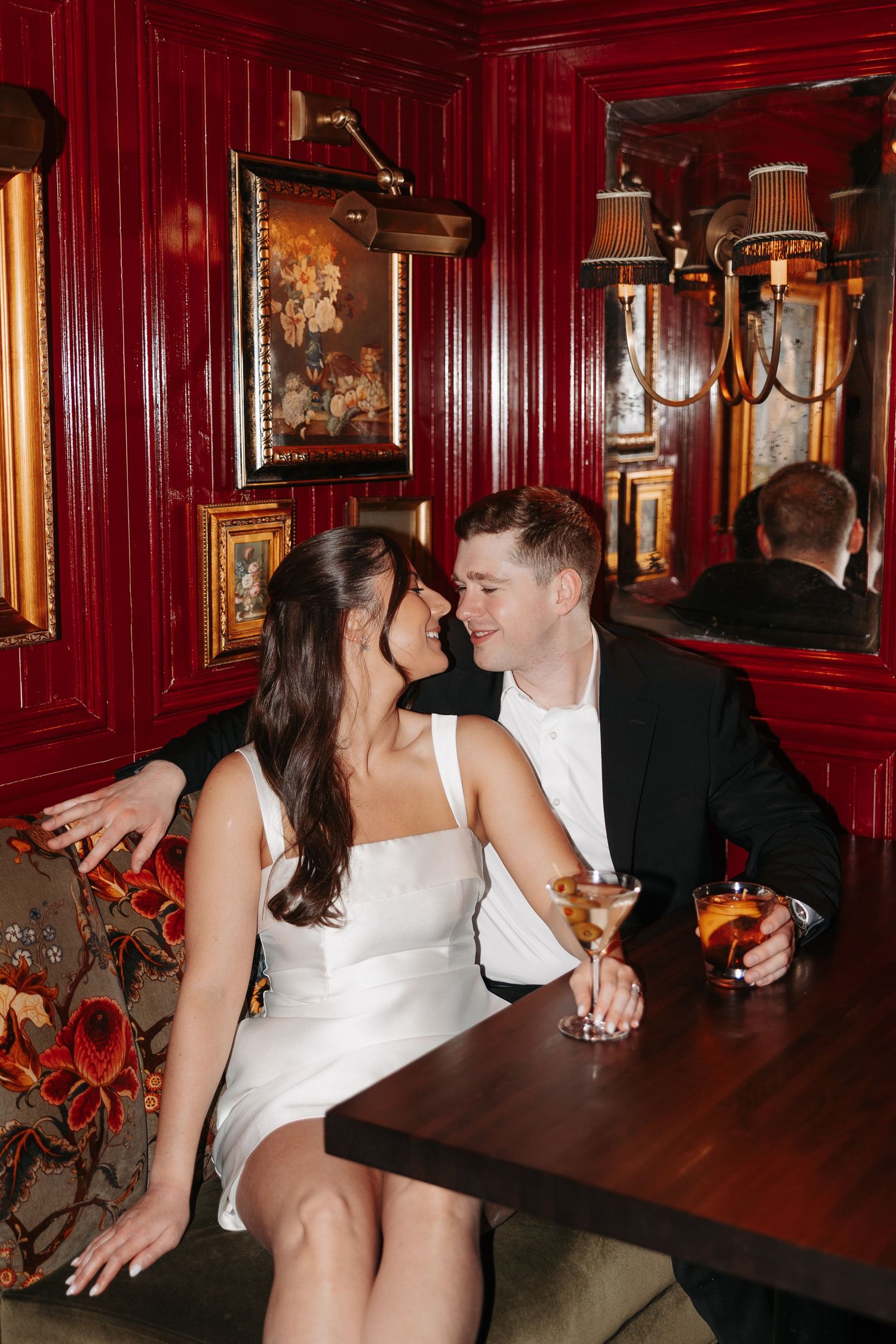 A romantic couple shares an intimate moment at a vintage bar with red walls and ornate decor while enjoying cocktails.