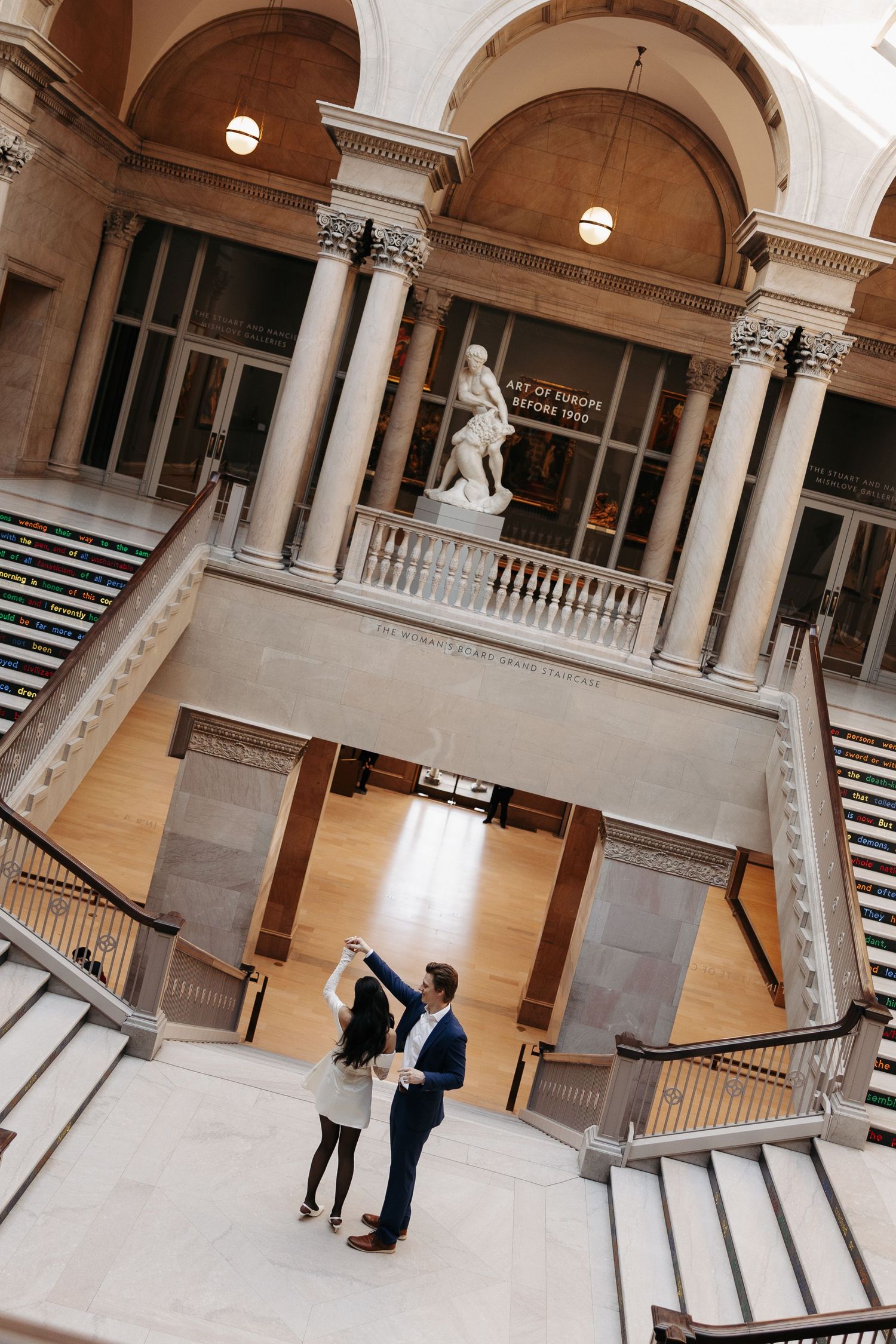 Two people stand beneath grand arched columns and a classical statue in an elegant interior stairwell with marble steps.