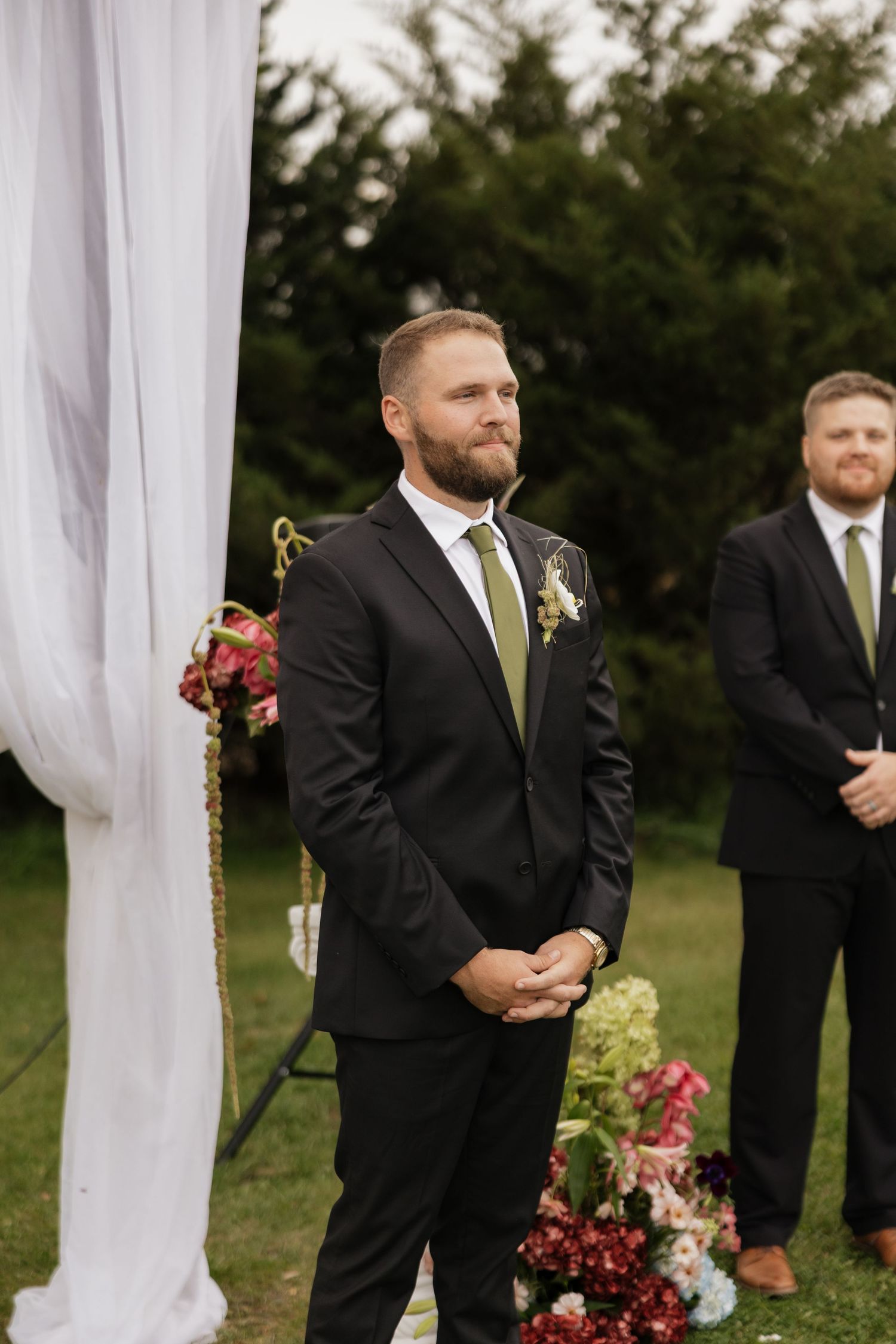 Bearded groom in a black suit and green tie stands at an outdoor wedding ceremony beside white drapes and pink floral arrangements.