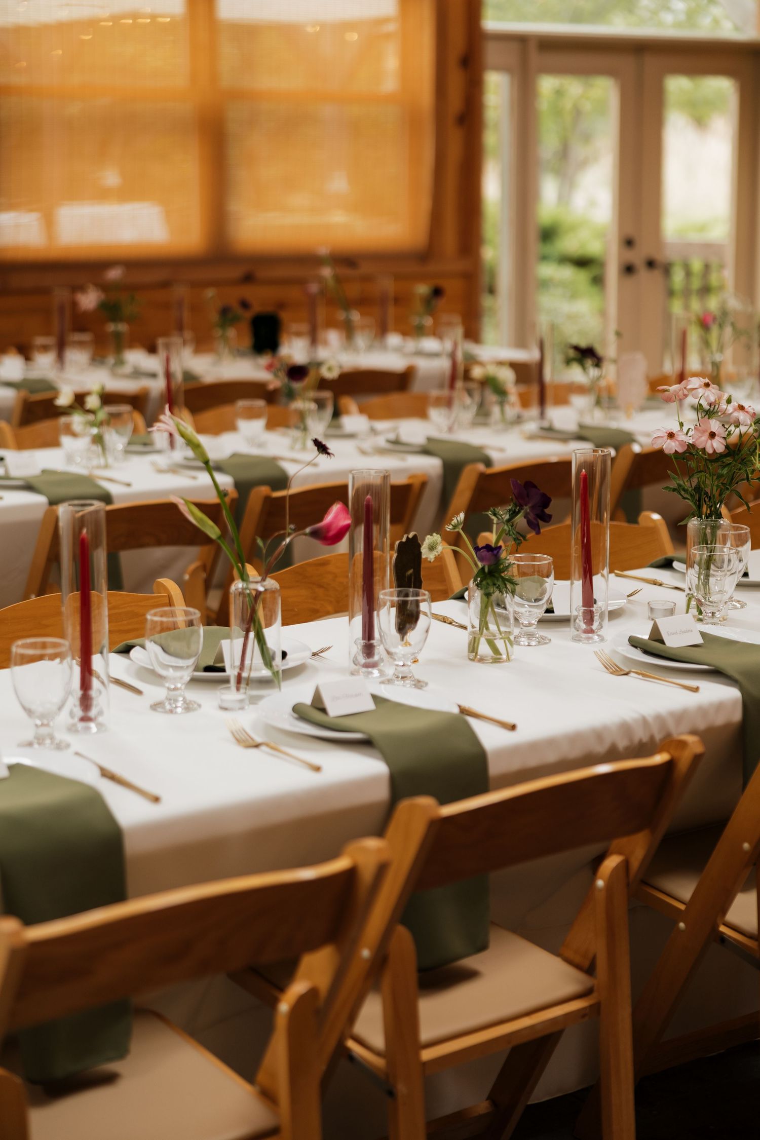 Elegant wedding reception tables with olive napkins, red candles, and wildflower centerpieces beside tall windows in a rustic venue.