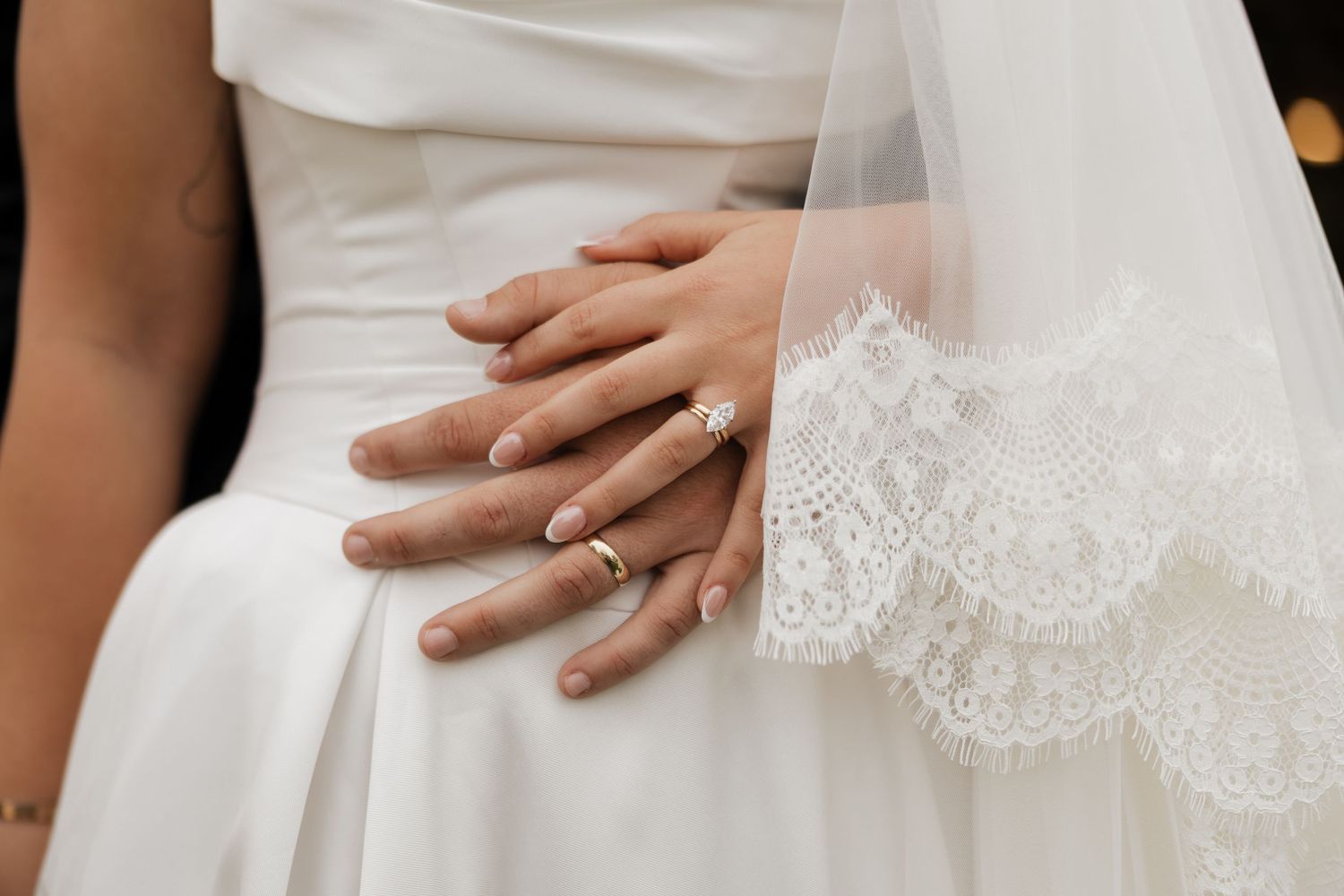 Bride's hand with a diamond ring resting on the groom's hand with a gold band, beside a white wedding dress and lace-trimmed veil.