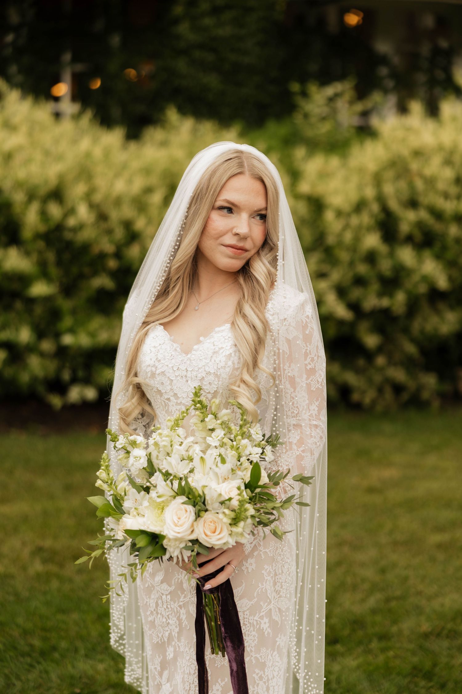 Bride in a lace wedding gown and long beaded veil holding a white rose bouquet with greenery in a sunlit garden.
