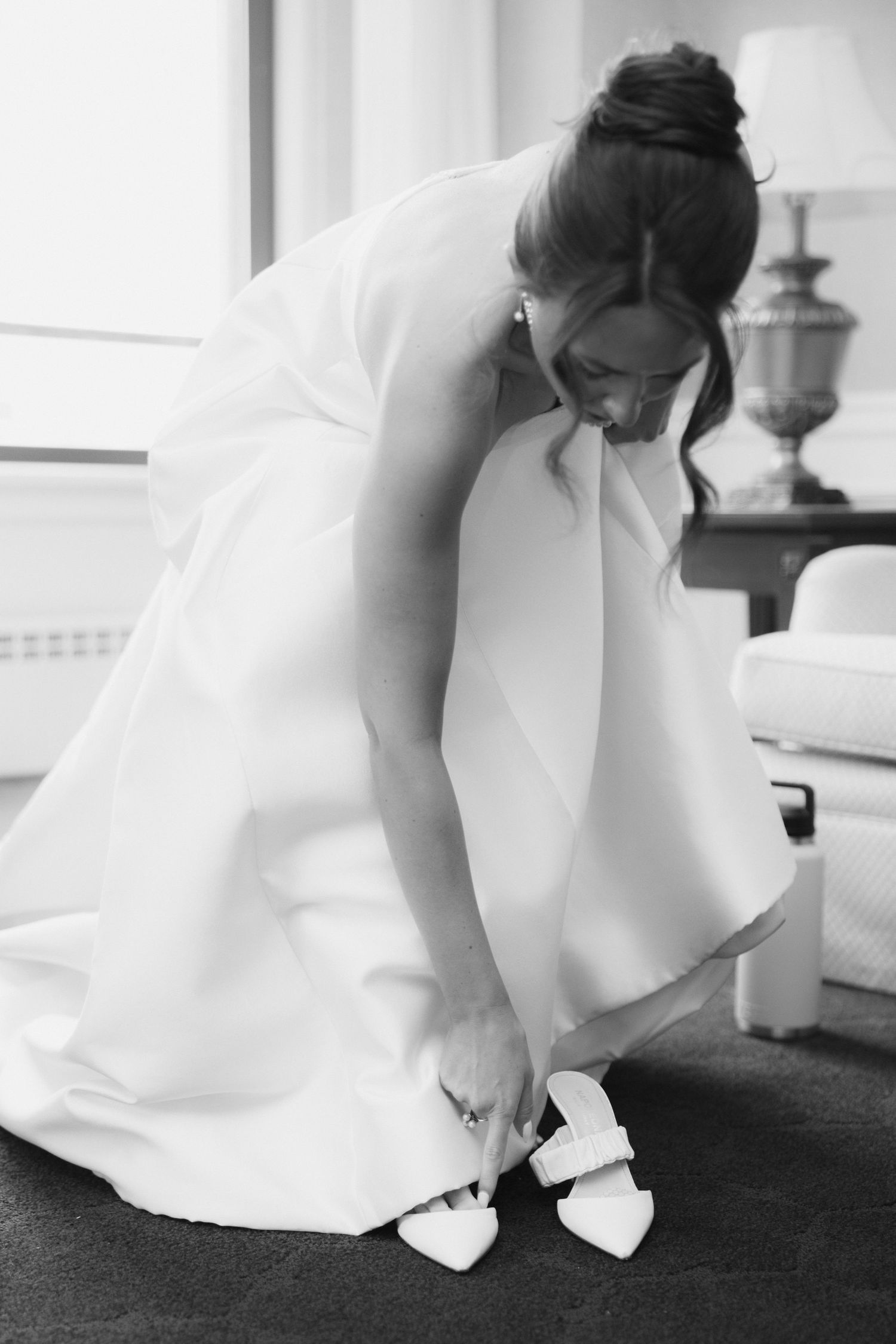 Bride in a white strapless gown with an updo bends down to slip on pointed white bridal heels in a softly lit room, black and white.