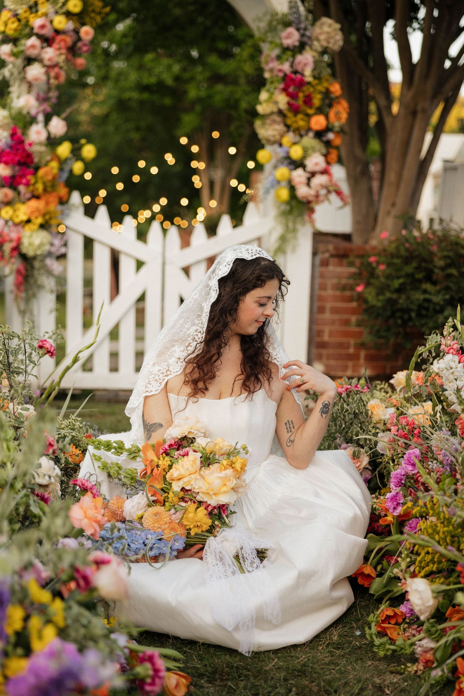 Bride in white gown and veil sits among colorful flowers by a white gate with string lights and floral backdrop.