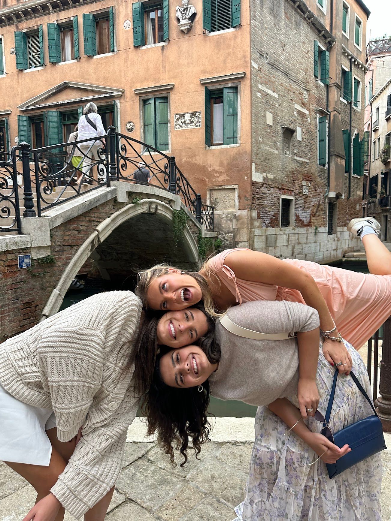 Three friends pose playfully on a canal bridge in Venice, Italy, with historic buildings and waterways in the background.