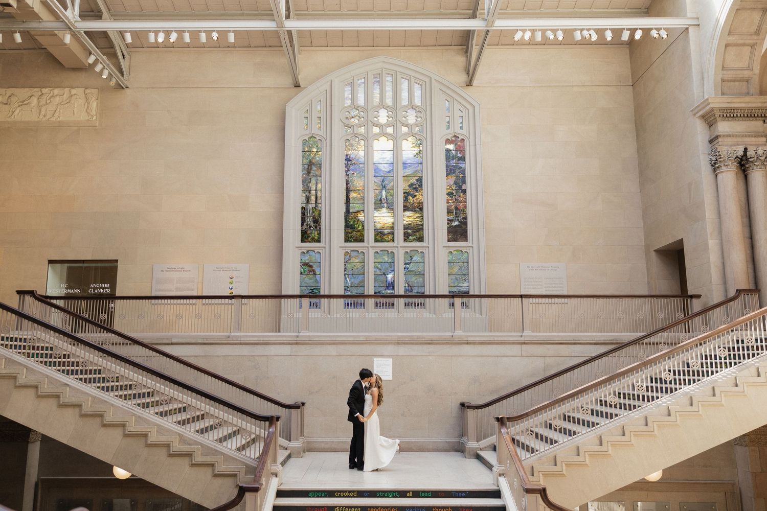 Bride and groom share a kiss on grand staircase beneath stunning stained glass window in elegant wedding venue.