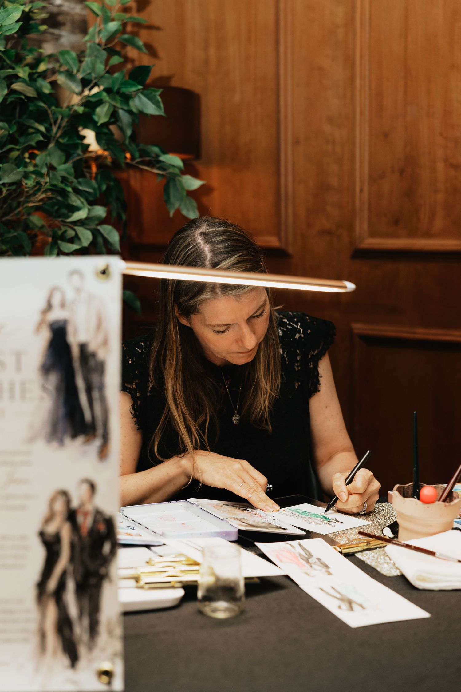 Artist sketching fashion illustrations at a desk with papers and art supplies at an elegant event.