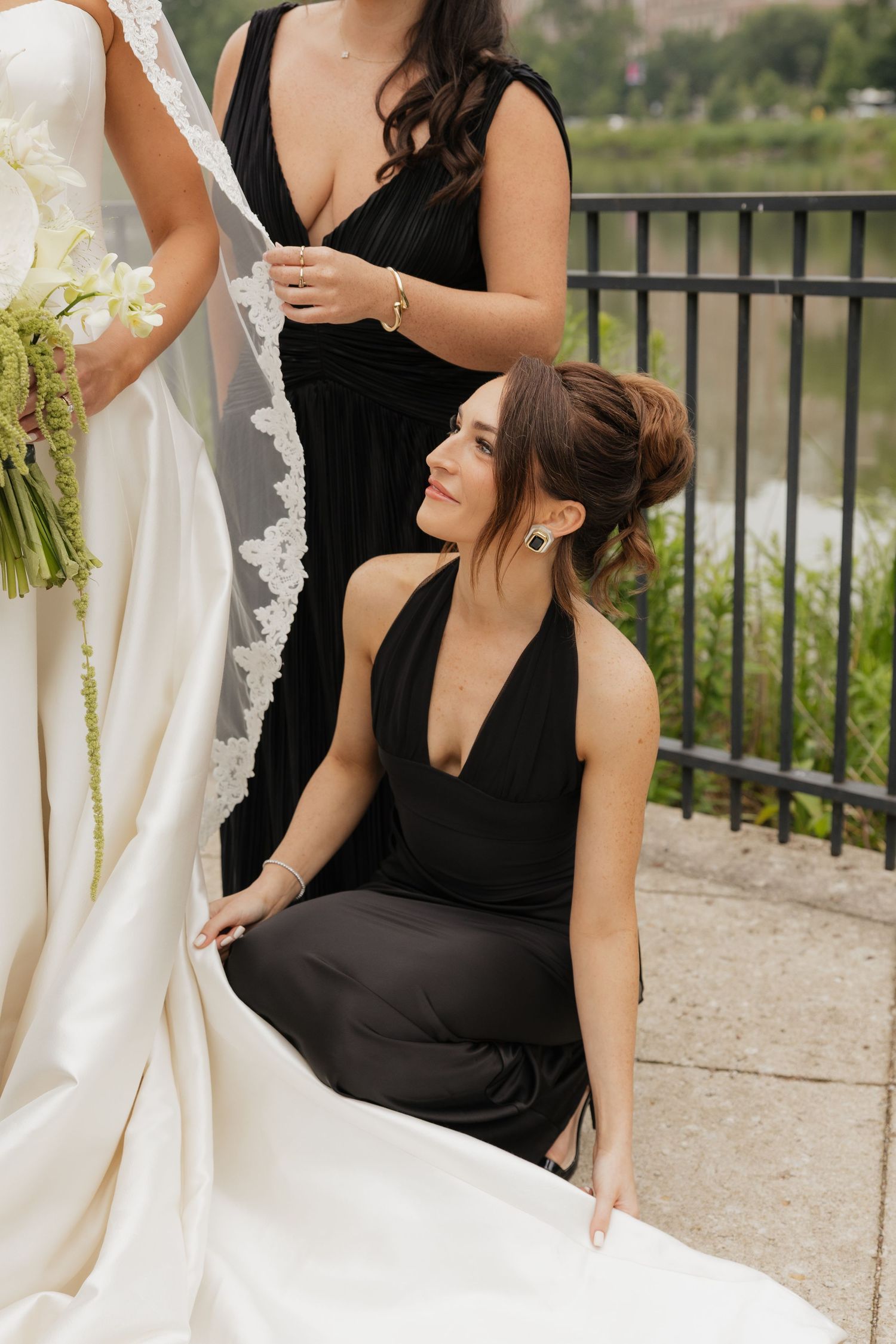 Two bridesmaids in black dresses assist a bride in a white gown outdoors near a railing.
