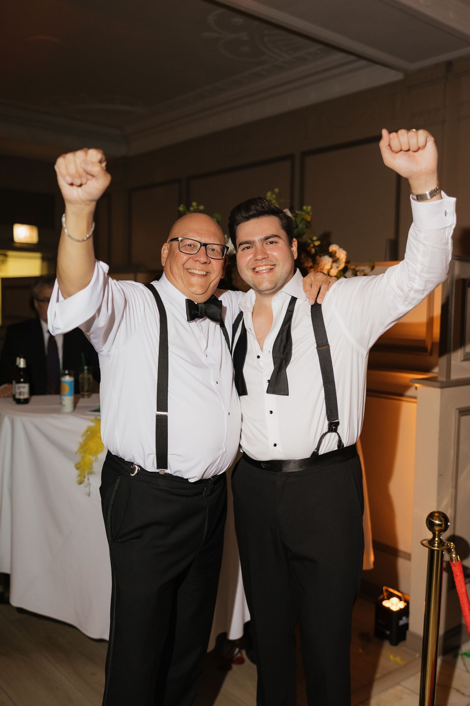 Two men in tuxedo shirts and suspenders celebrate with raised fists at a formal wedding reception event.