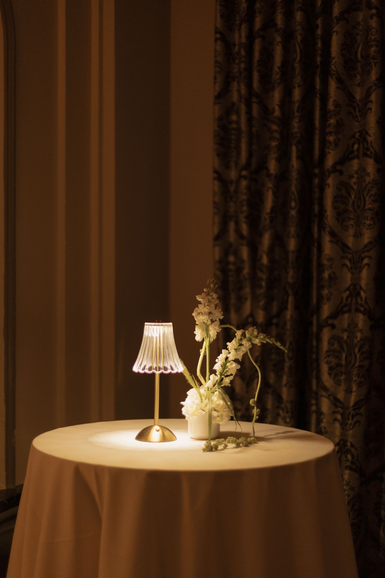 Elegant table setting with glowing lamp and white flowers on draped table against ornate curtains.