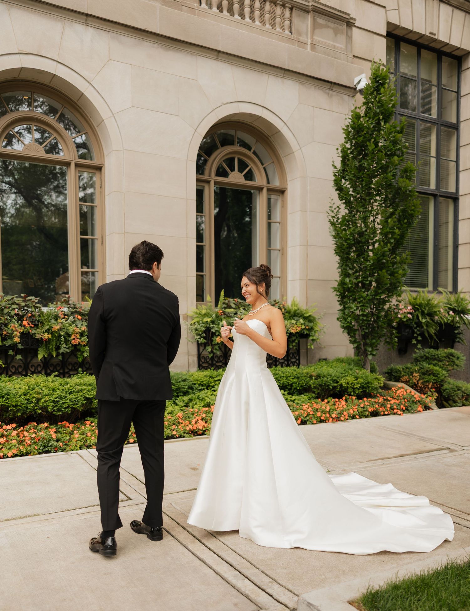 Bride in white gown reads vows to groom in black tuxedo outside elegant stone building with lush gardens.