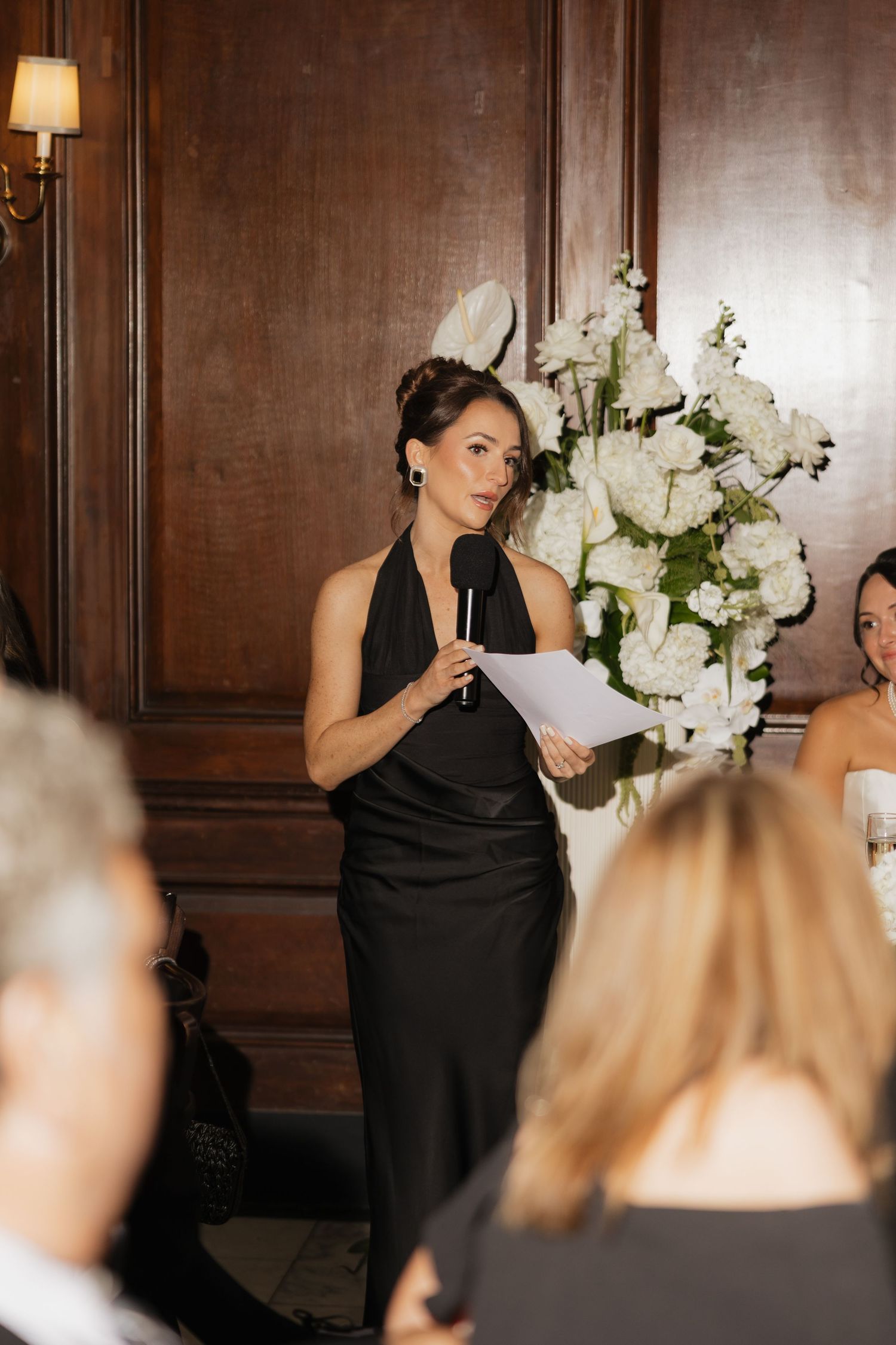 Woman in black halter dress gives speech at elegant event, holding microphone and papers near white floral arrangement.