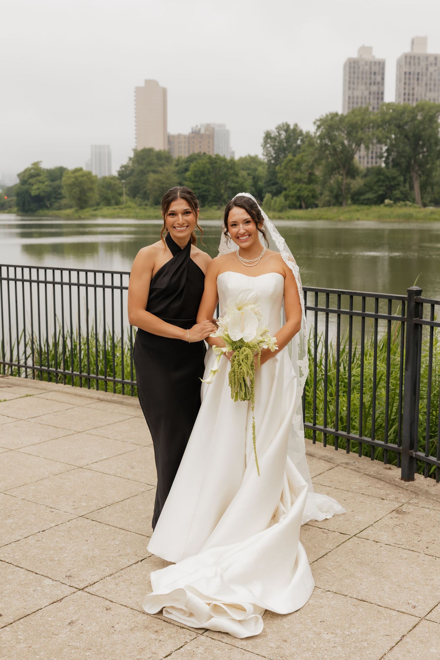 Bride in white gown with veil holds bouquet beside bridesmaid in black dress, posing by a lakeside railing with city skyline.