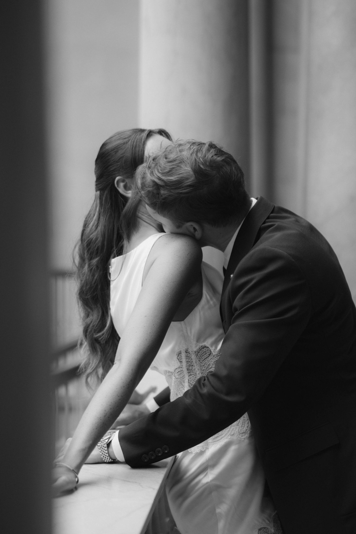 Black and white photo of a couple sharing a romantic kiss, bride in white dress and groom in suit near columns.