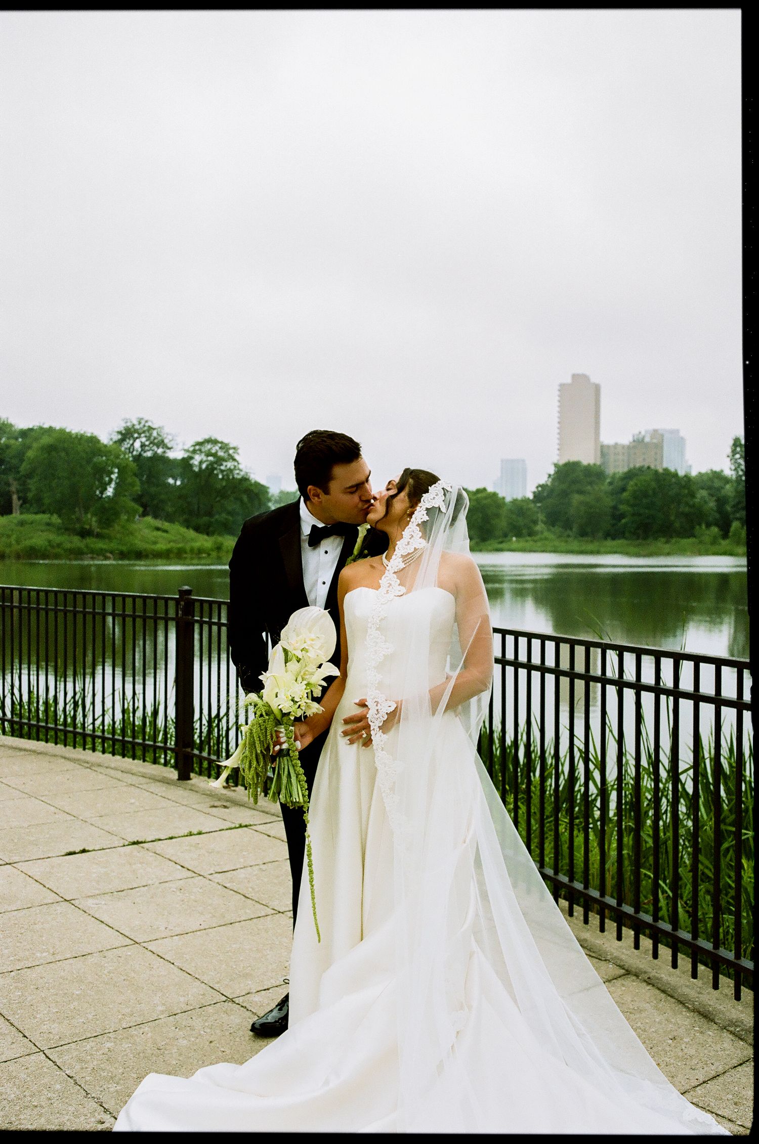 Bride and groom share romantic kiss by lakeside with city skyline backdrop on wedding day.