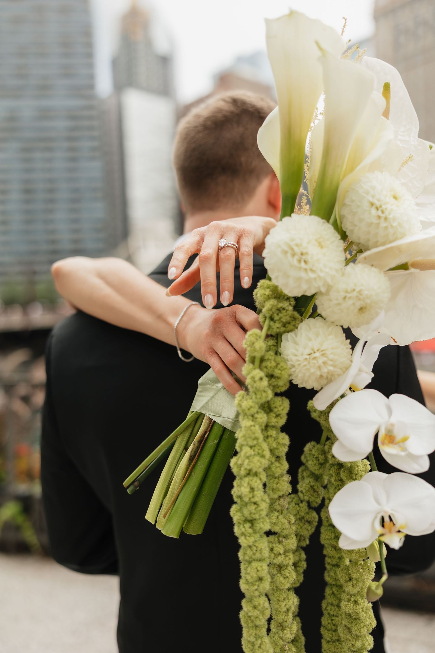 Bride holding white calla lily and orchid bouquet while embracing groom on rooftop with city skyline backdrop.