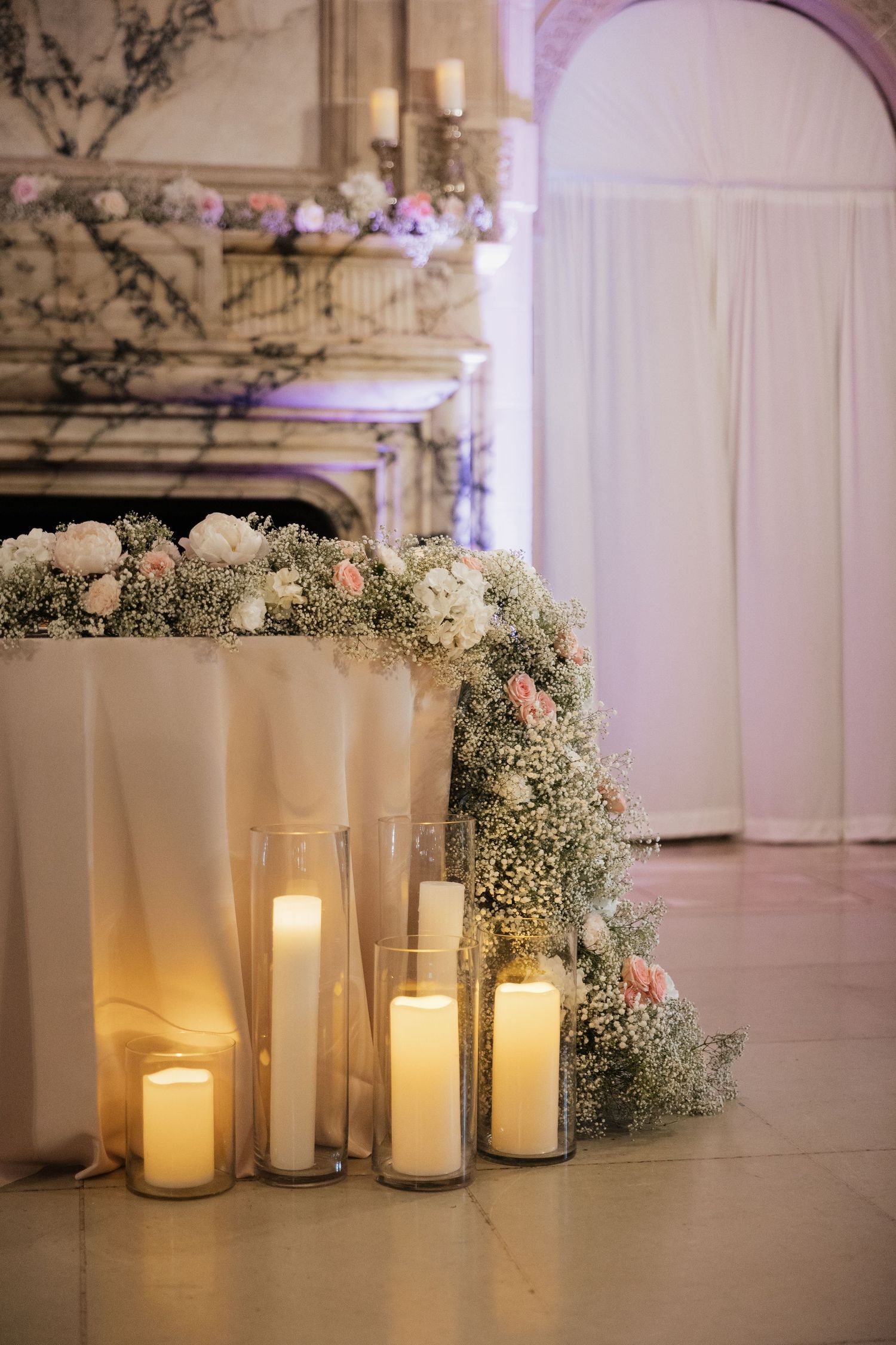 Elegant wedding table decorated with white candles in glass holders, pink roses, and baby's breath flowers near a marble fireplace.