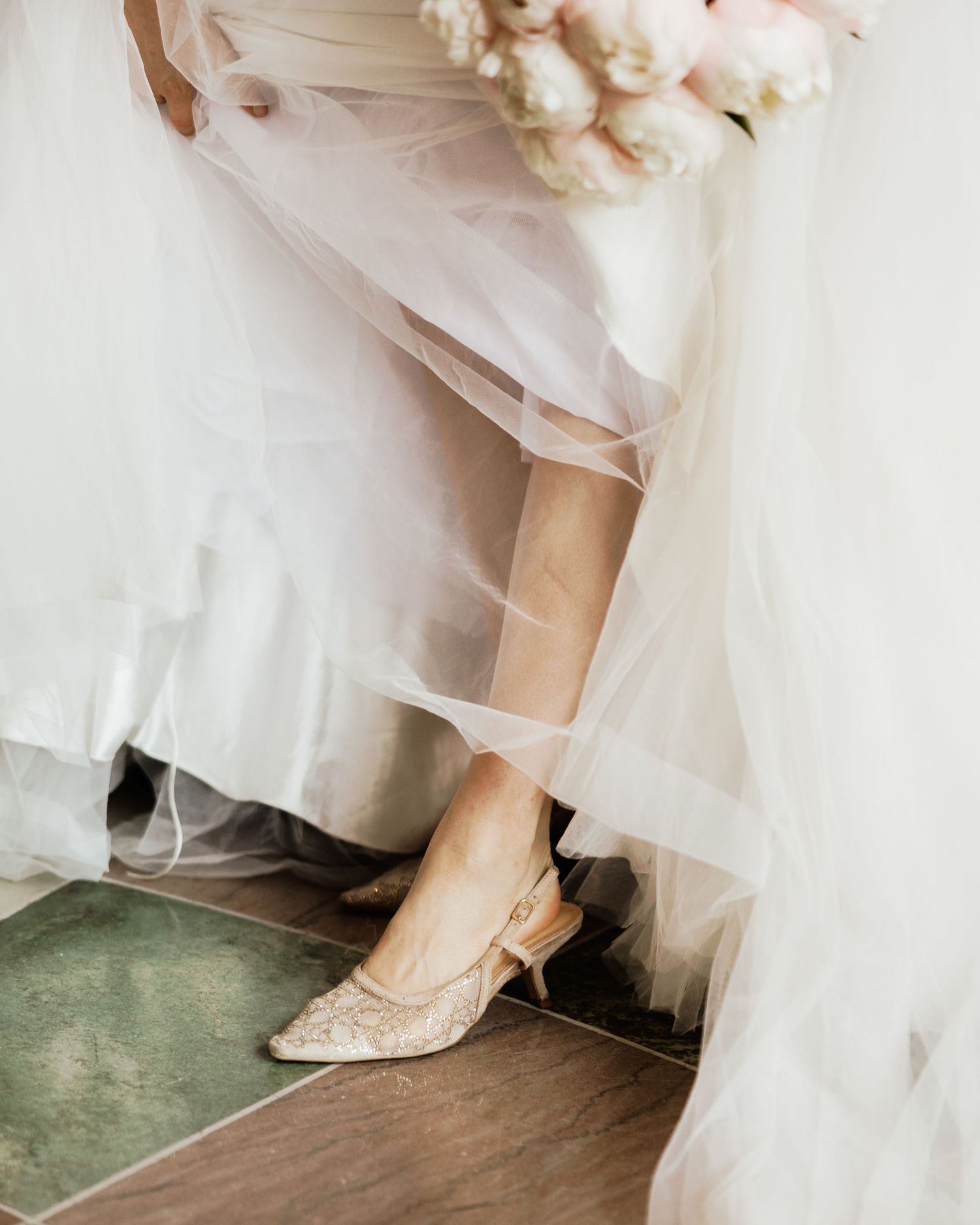 Bride in white tulle wedding gown wearing elegant lace kitten heel slingback shoes, holding a soft pink peony bouquet.