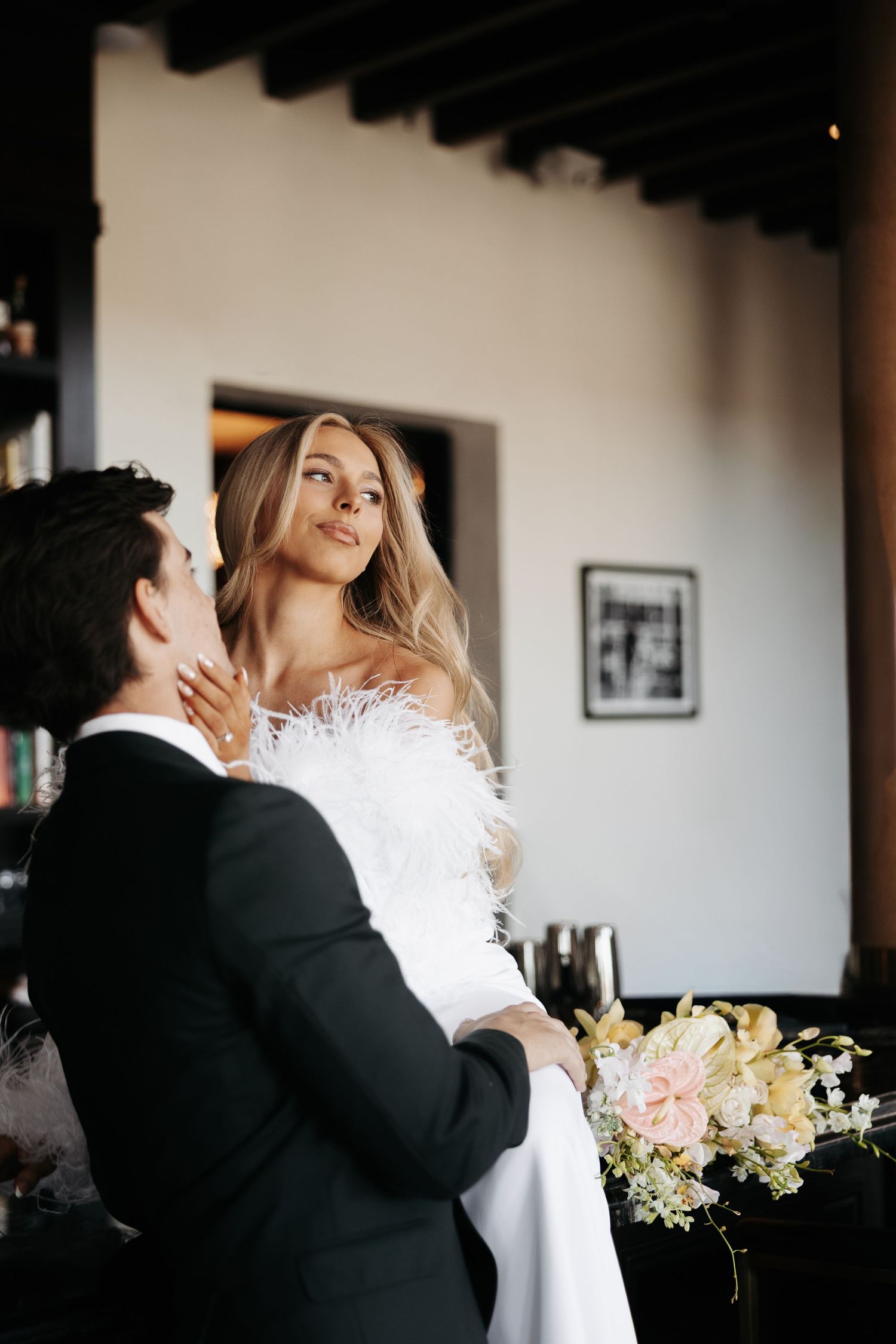 Romantic wedding couple embraces in an intimate moment by a window with soft natural light and floral arrangements.