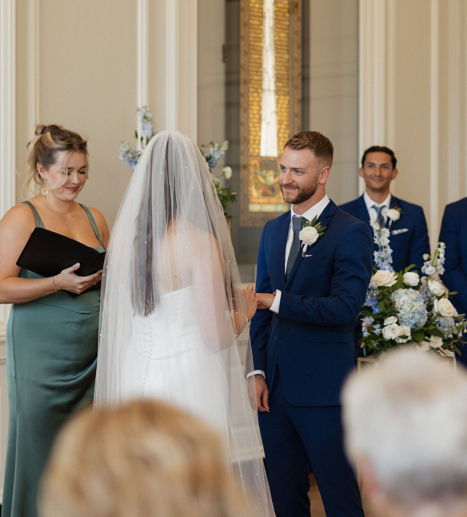 Wedding party members in navy suits and green dresses gather during ceremony in elegant ballroom setting.