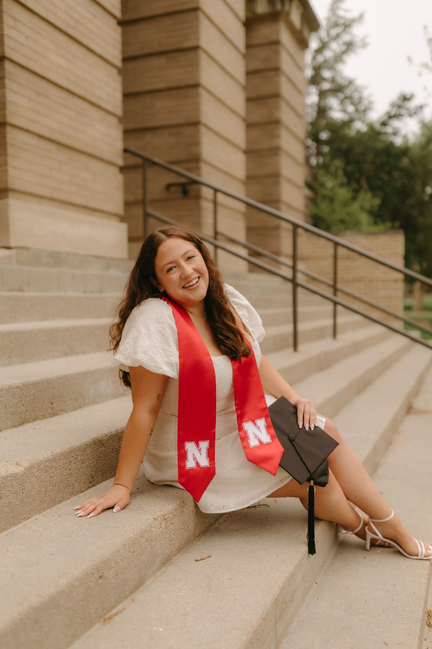 Graduate in red stole and white dress sits on concrete steps during graduation photoshoot at university.