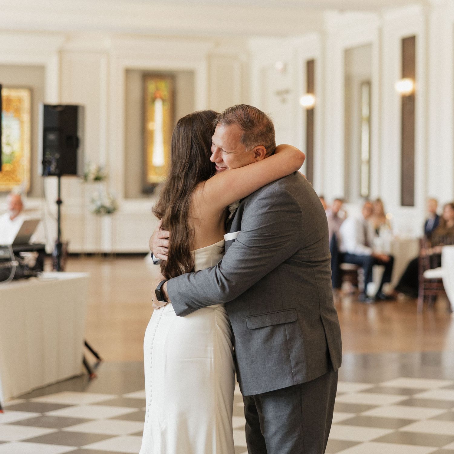 A couple shares their first dance in an elegant ballroom with white decor and guests seated in the background.