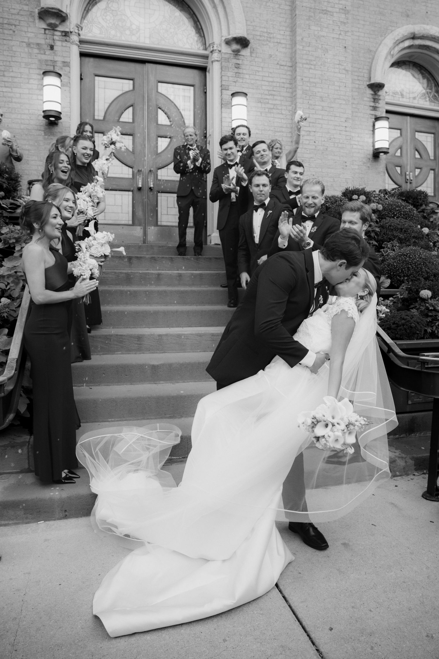 Groom dips bride for a dramatic kiss on church steps as joyful bridesmaids and groomsmen cheer in this black and white wedding photo.