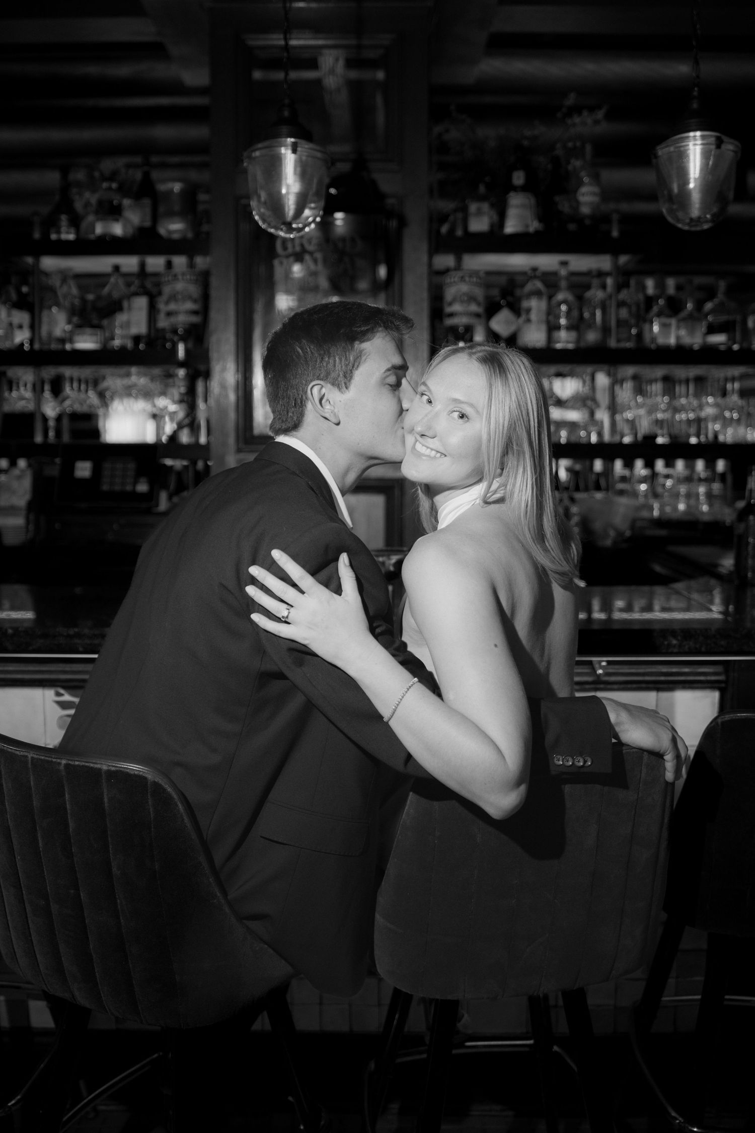 Black and white photo of a man in a suit kissing a smiling woman's cheek as they sit at a bar, with a ring visible on her hand.