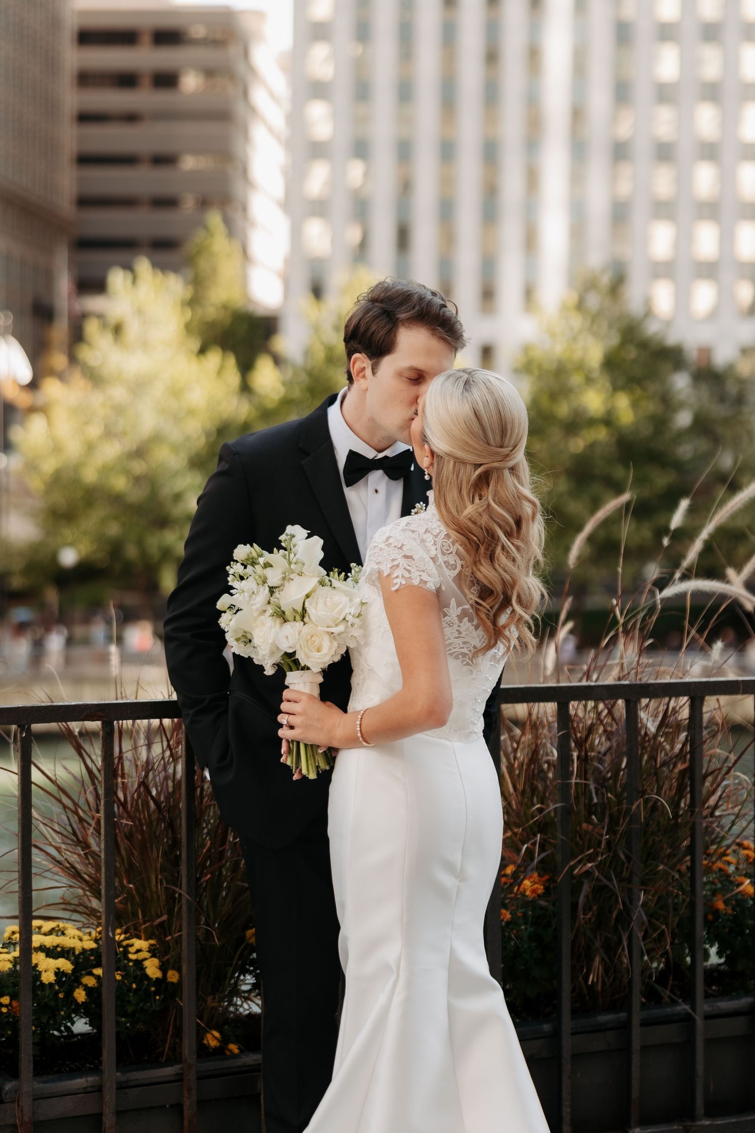 Bride in a lace mermaid wedding gown holding a white bouquet kisses her groom in a black tuxedo and bow tie on a city balcony.