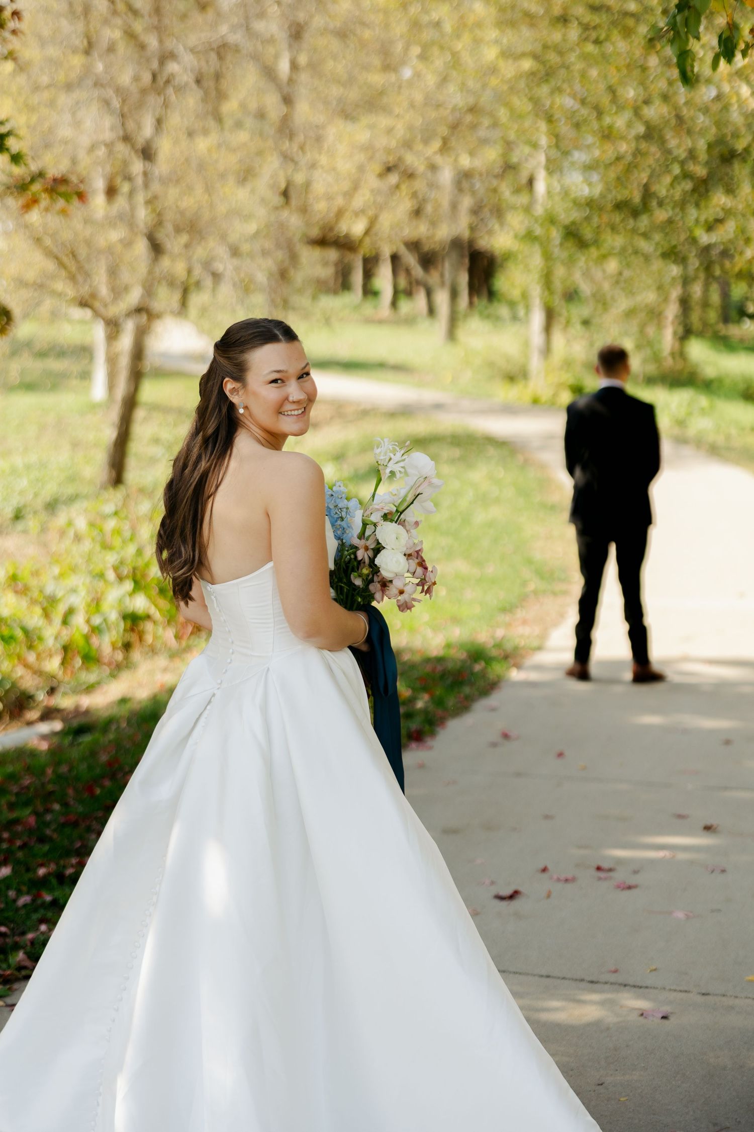Smiling bride in a strapless white gown holds a bouquet and looks back over her shoulder while the groom stands facing away on a park path.