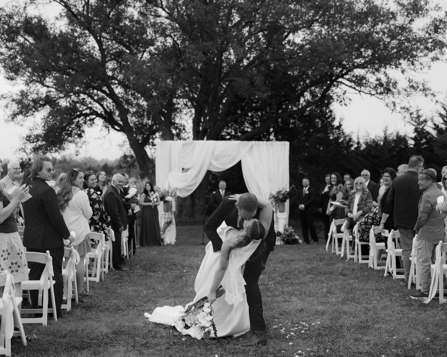 Groom dips bride in a romantic kiss at an outdoor wedding ceremony as seated guests watch beneath large trees and a white draped altar.