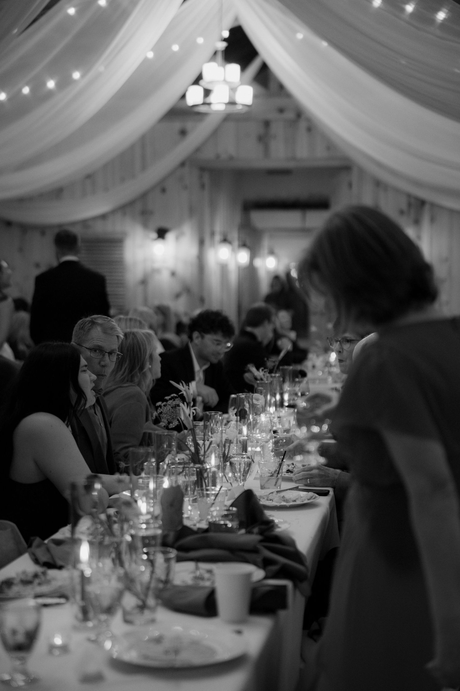 Black and white photo of wedding guests seated at a long candlelit banquet table in a rustic barn with draped fabric and string lights.