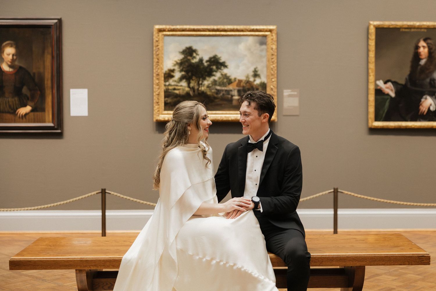 A couple in wedding attire sits on a wooden bench in an art gallery with classic paintings on display.