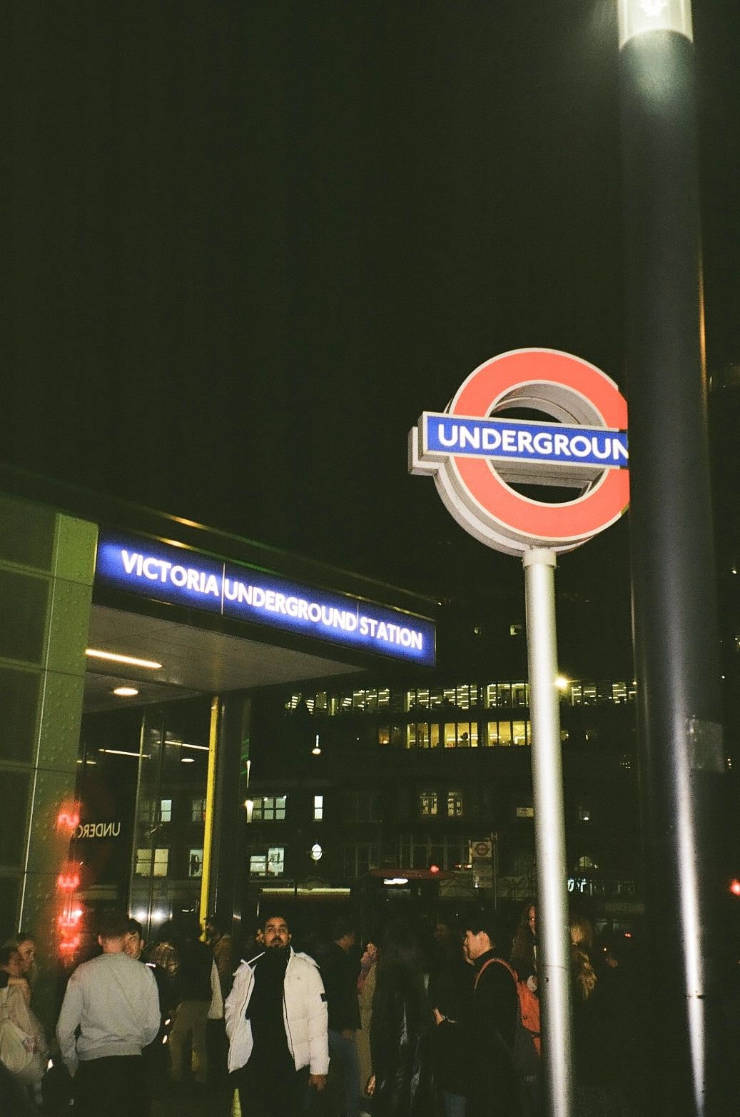 Nighttime view of Victoria Underground station entrance in London with illuminated signs and commuters.