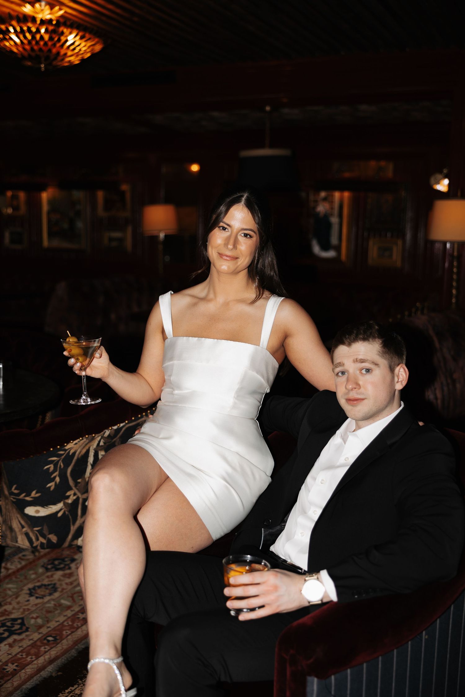 A couple in formal attire poses together on a dark couch at an evening event.