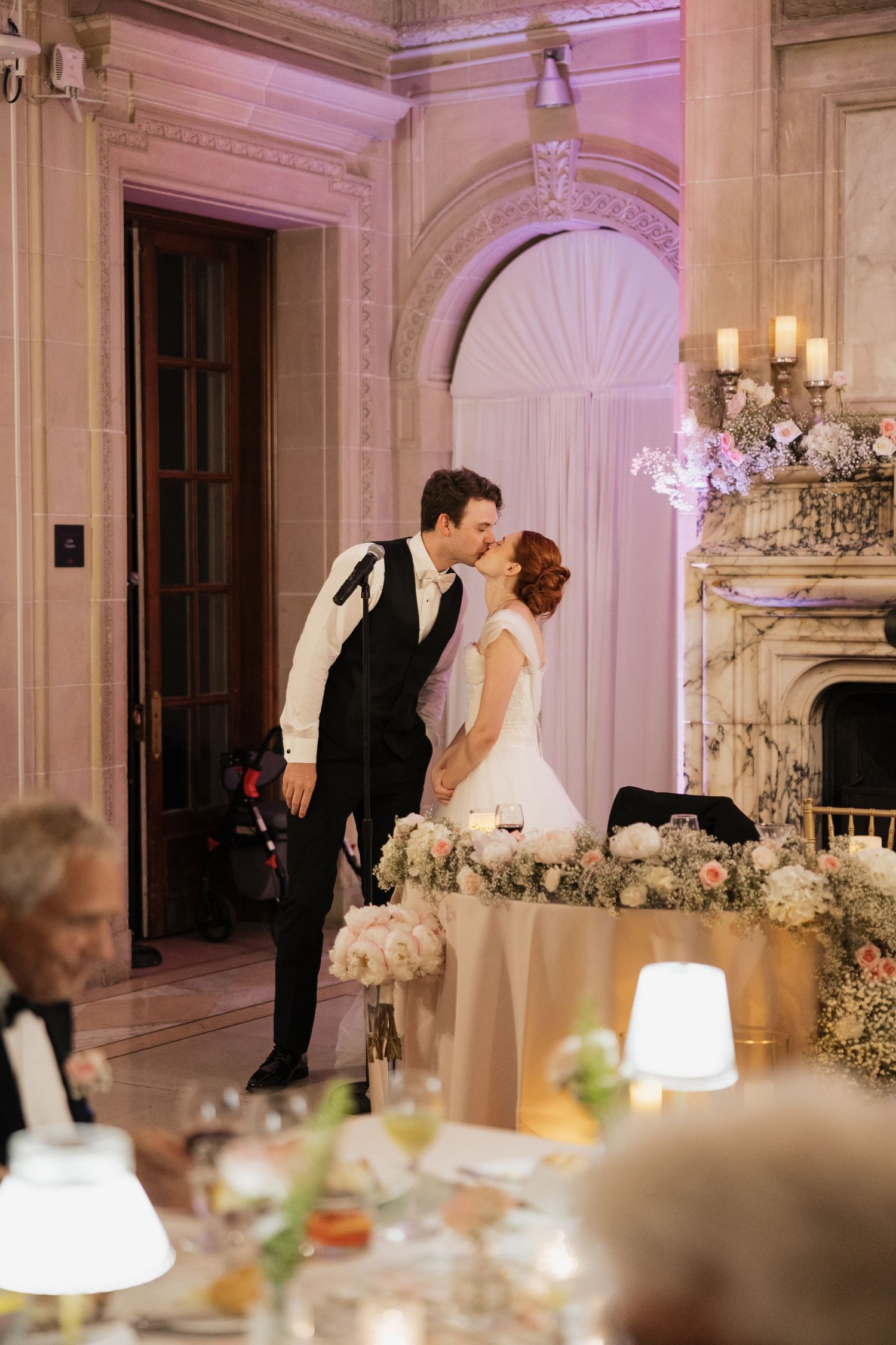 Bride and groom share a romantic kiss at their elegant wedding reception beside a floral centerpiece and grand fireplace.