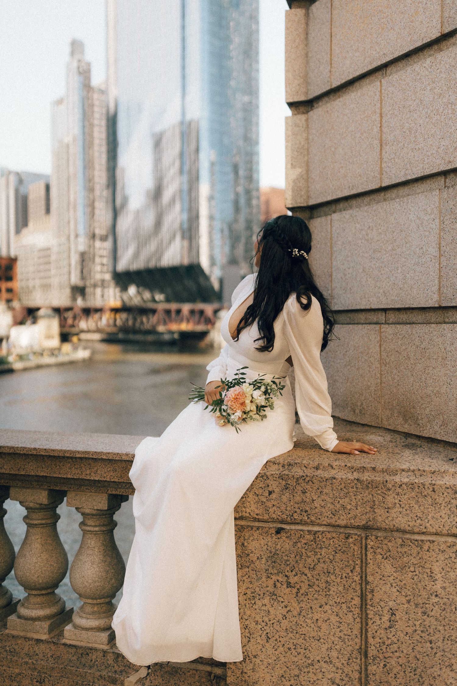 A person in a flowing white dress sits elegantly on a stone balustrade with a city skyline in the background.
