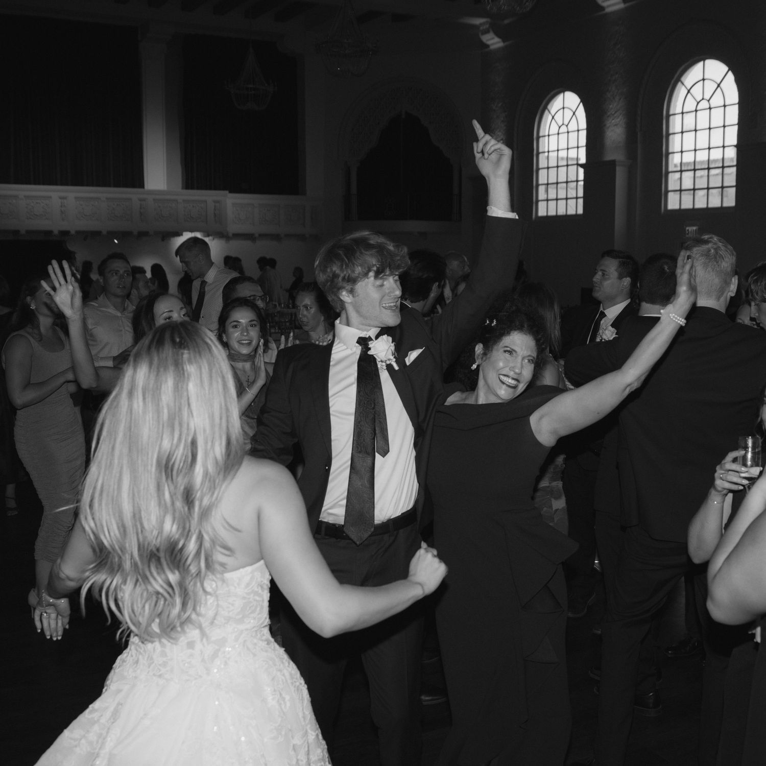 Wedding guests dancing and celebrating at a dark reception venue with arched windows.