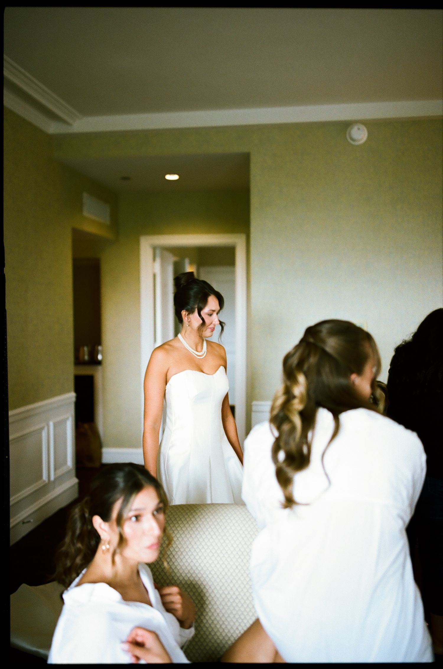 Bride in white strapless gown and pearl necklace smiles while bridesmaids help her prepare in elegant hotel room.