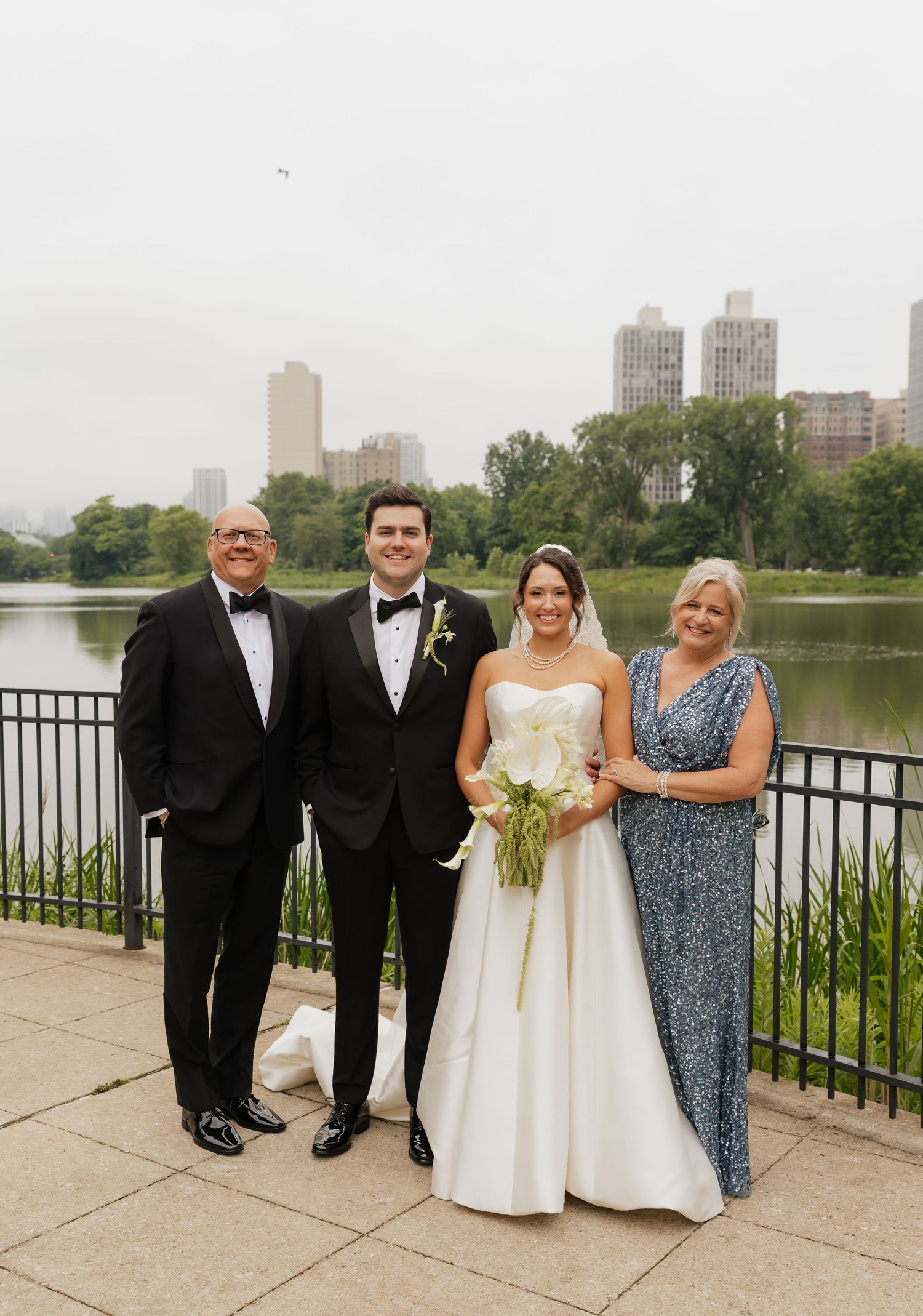 Wedding party of four posing by a lakeside park with city skyline backdrop, bride in white gown holding calla lilies.