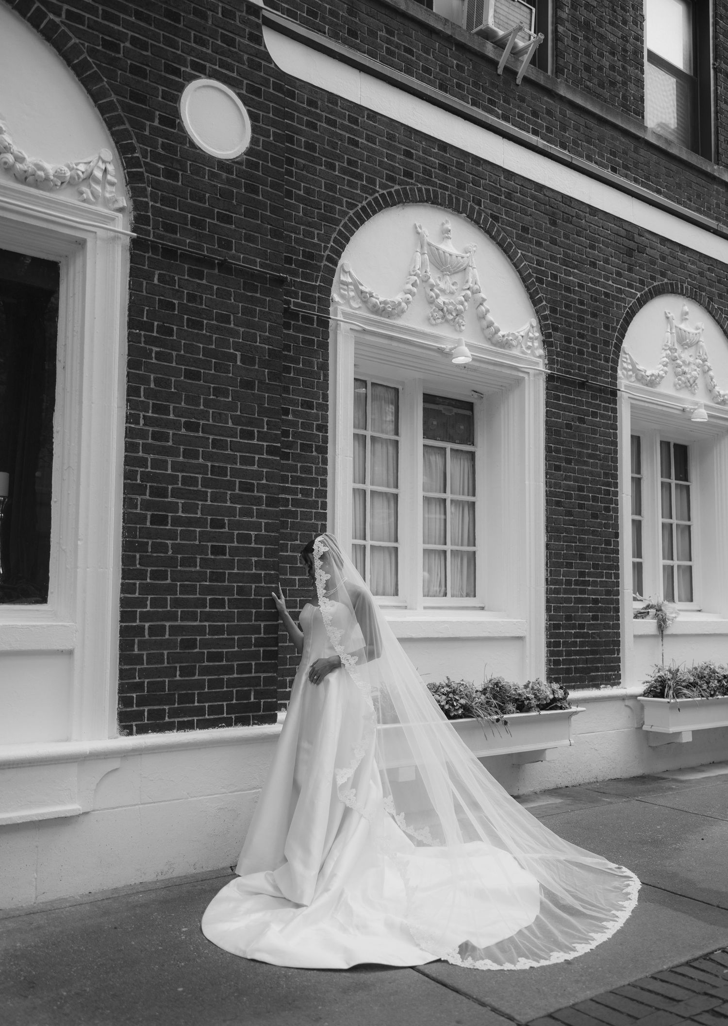 Black and white photo of a bride in a flowing gown and veil posing outside an elegant brick building.